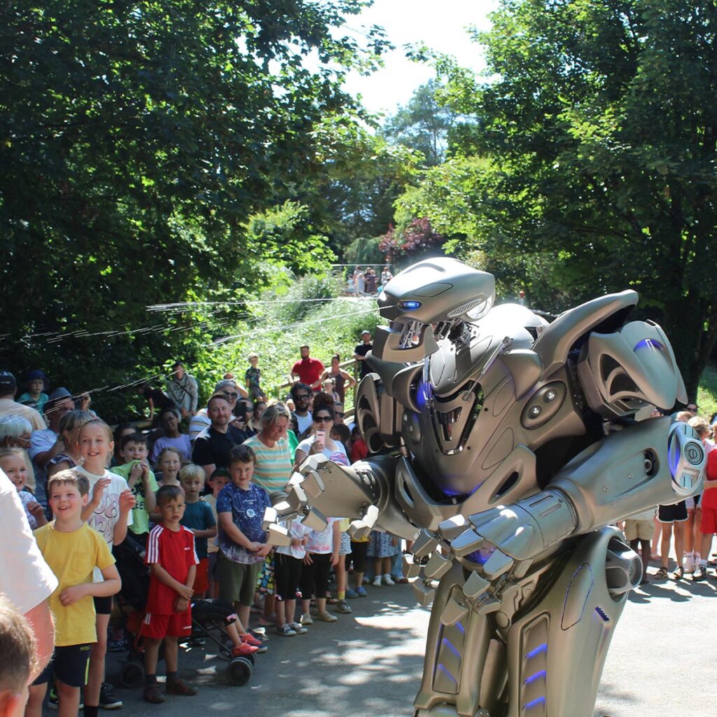 Titan the Robot squirts water over a crowd of children at Woodlands Family Theme Park, surrounded by tall green trees in the sunshine.